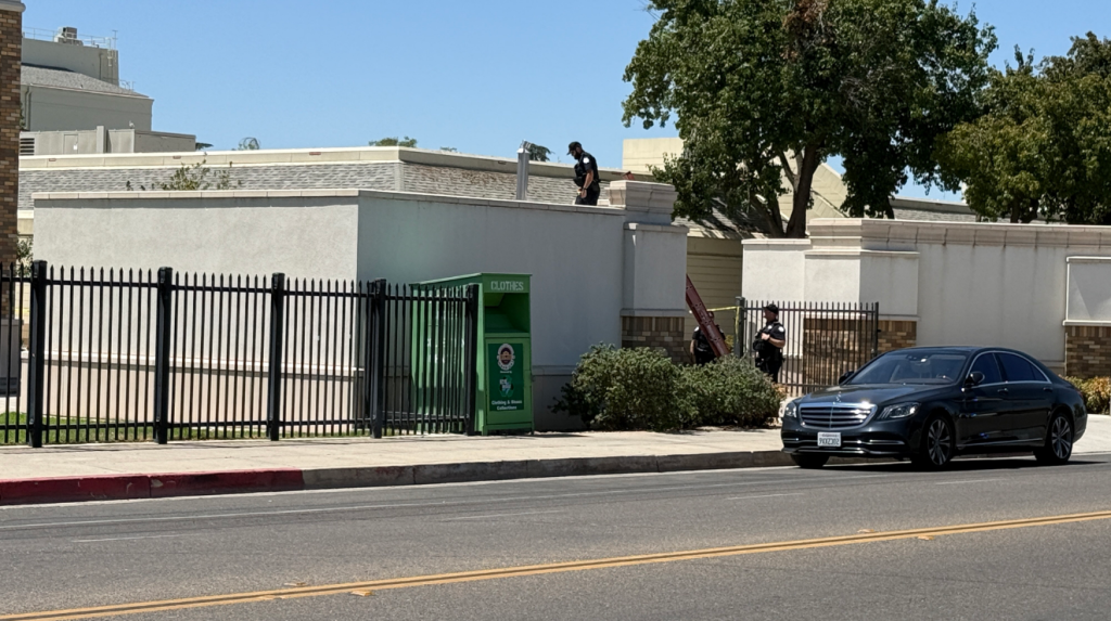 A police officer standing on the roof of a building at Fresno High School, with another officer and a black sedan on the street below.