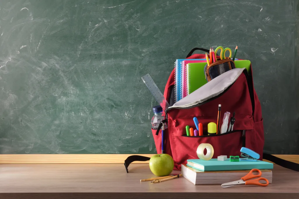 Red backpack filled with school supplies on a wooden table in front of a green chalkboard.