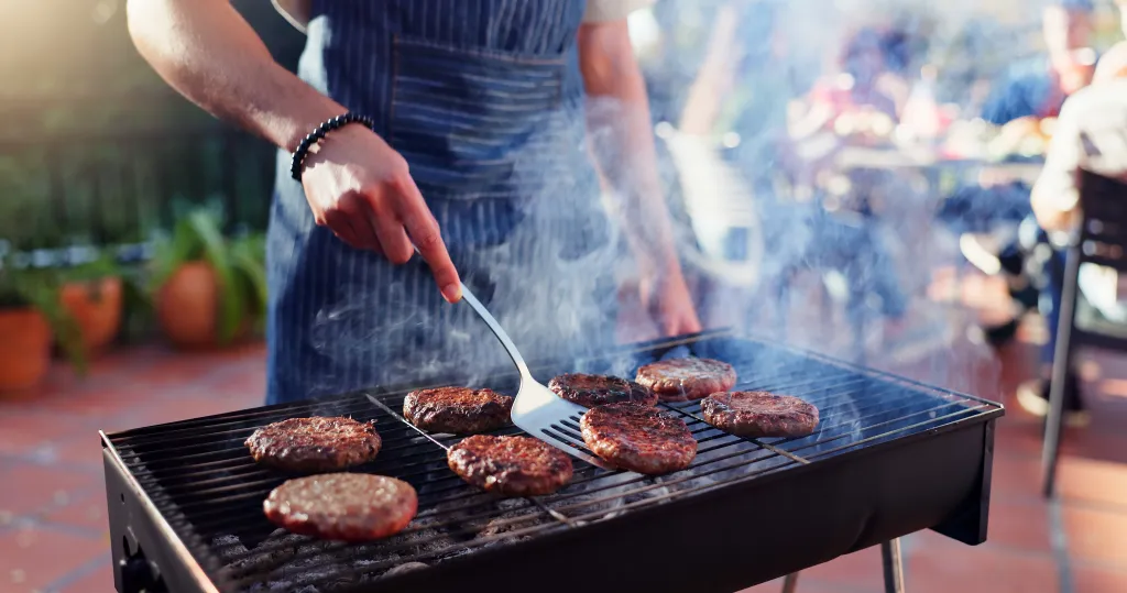 A man in an apron grilling beef patties on an outdoor barbecue.