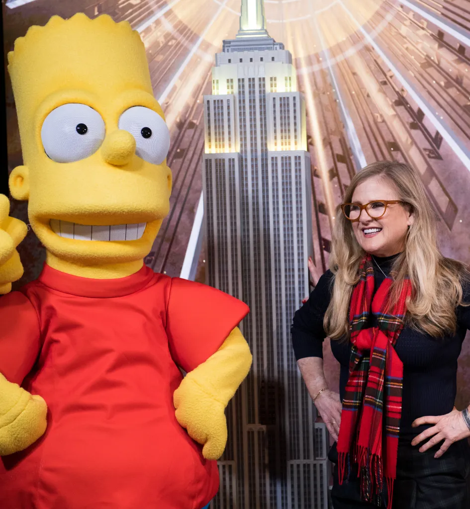 Nancy Cartwright, the voice of Bart Simpson, and a costumed Bart stand in front of a poster of the Empire State Building.