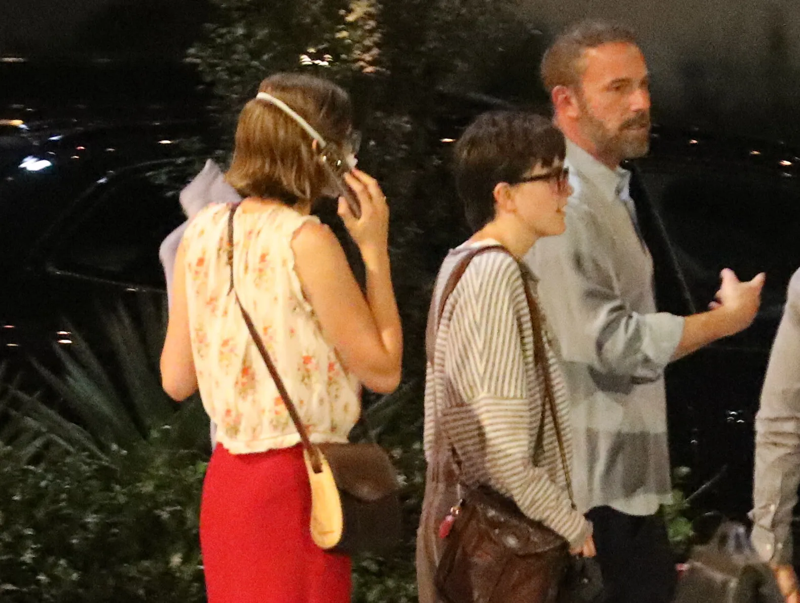 Ben Affleck, with a beard, walking outdoors at night with his two children, one of whom is masked.