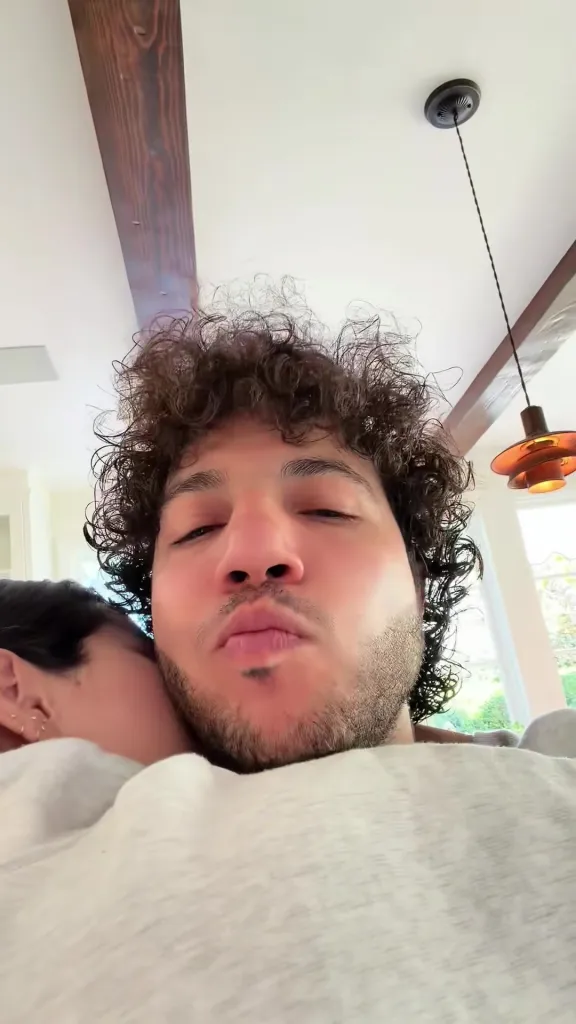Benny Blanco in a close-up selfie, making a kissing face, with a woman resting her head on his shoulder.
