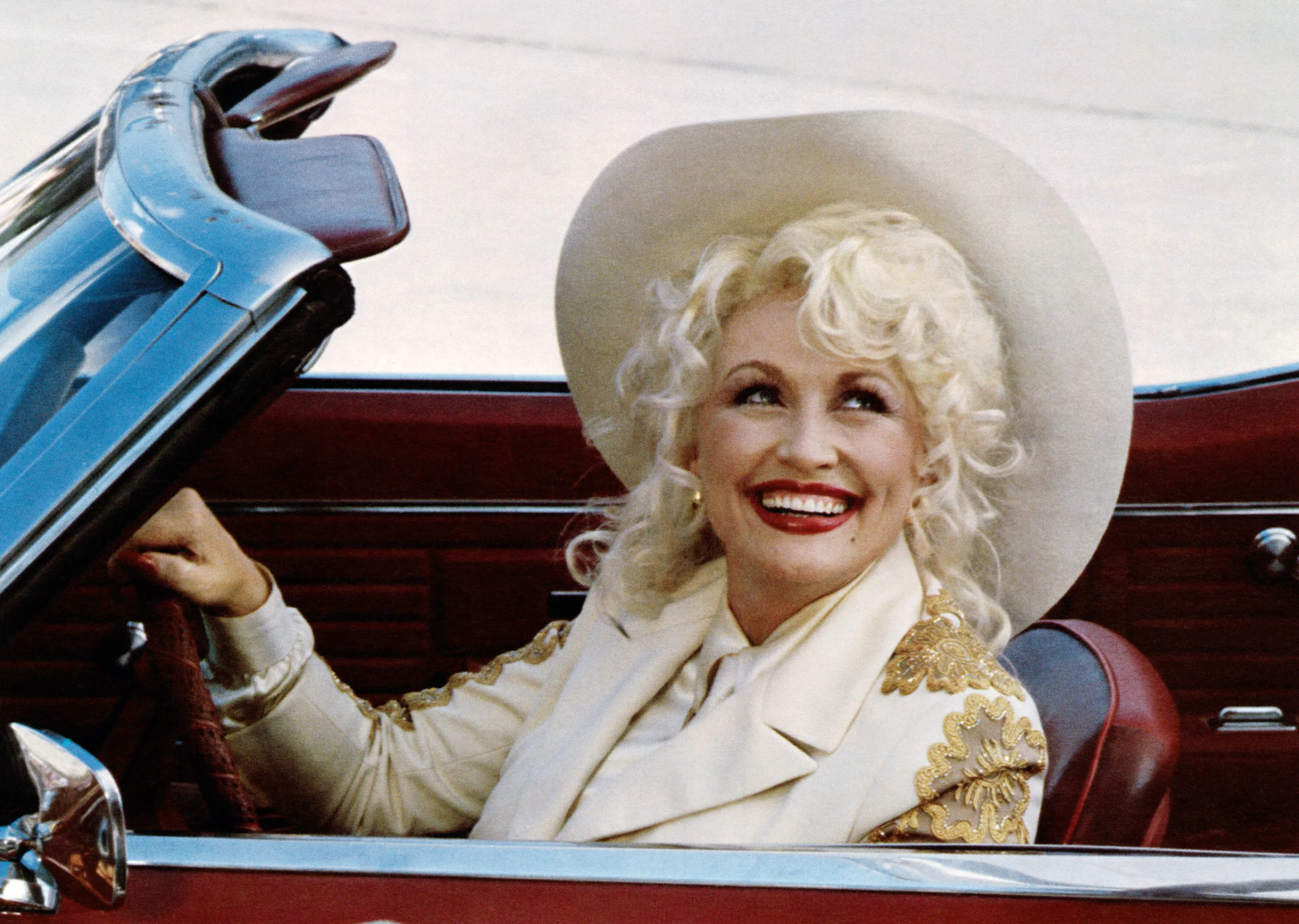 Dolly Parton in a convertible, wearing a white cowboy hat and embroidered jacket.