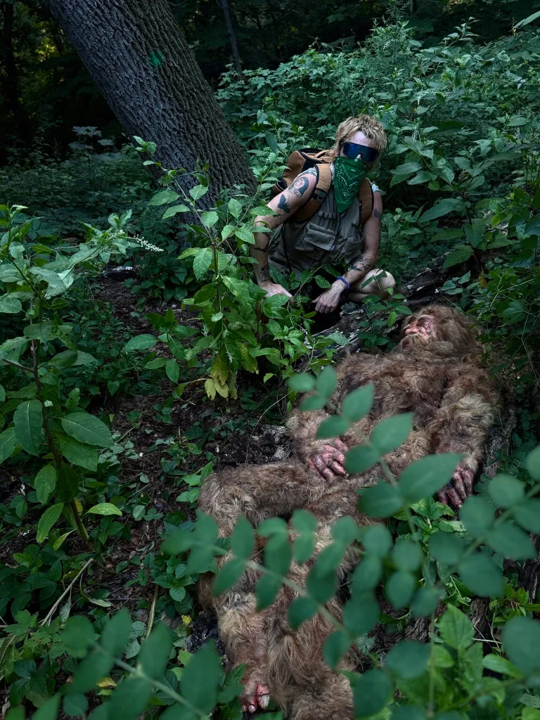 A hunter wearing a bandana and sunglasses crouches over a deceased Bigfoot in the woods.