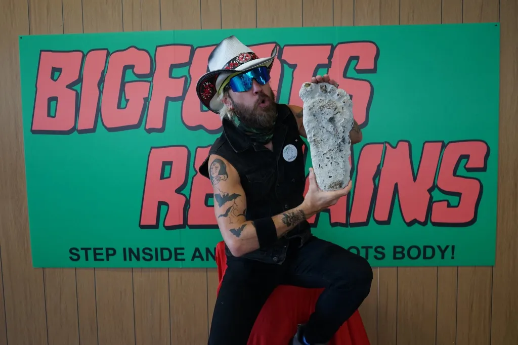 A man in a cowboy hat and sunglasses holds a large cast of a footprint, sitting in front of a green banner that reads 