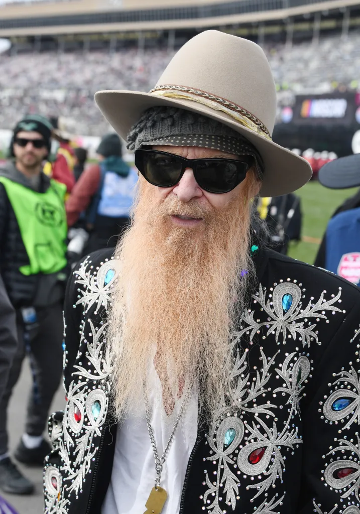 Billy Gibbons of ZZ Top in a black studded jacket, sunglasses, and a tan hat.