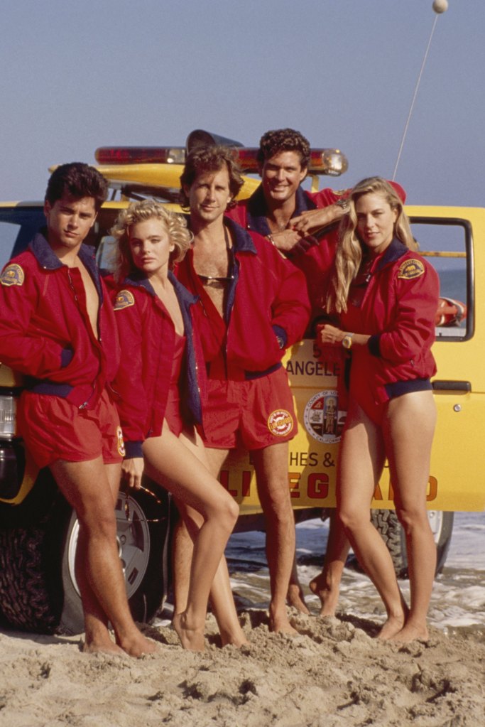 Billy Warlock, Erika Eleniak, Parker Stevenson, David Hasselhoff, and Shawn Weatherly pose on a beach with a lifeguard vehicle.