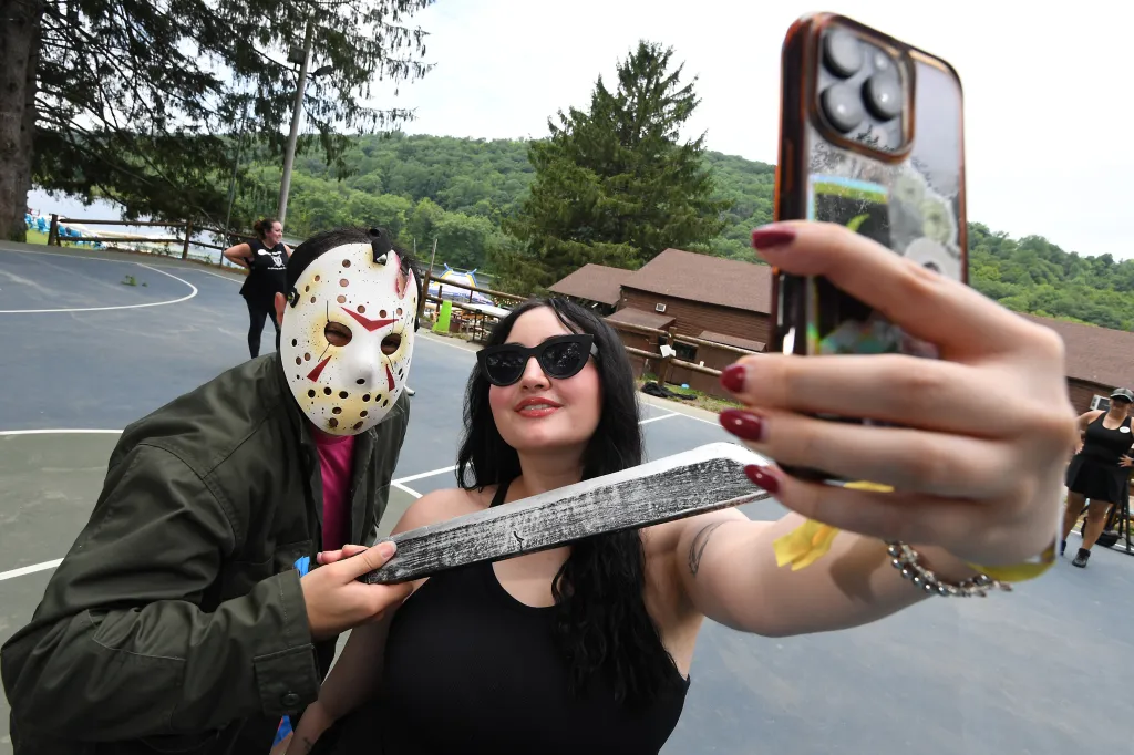 Marissa Matozzo (black hair/black top) taking a selfie with a person in a Jason mask holding a machete at Camp Getaway.