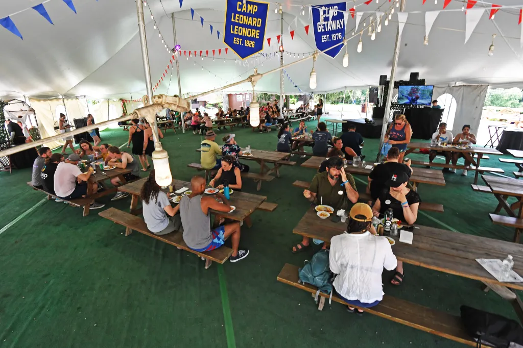 People eating and socializing at picnic tables under a large tent with banners for 