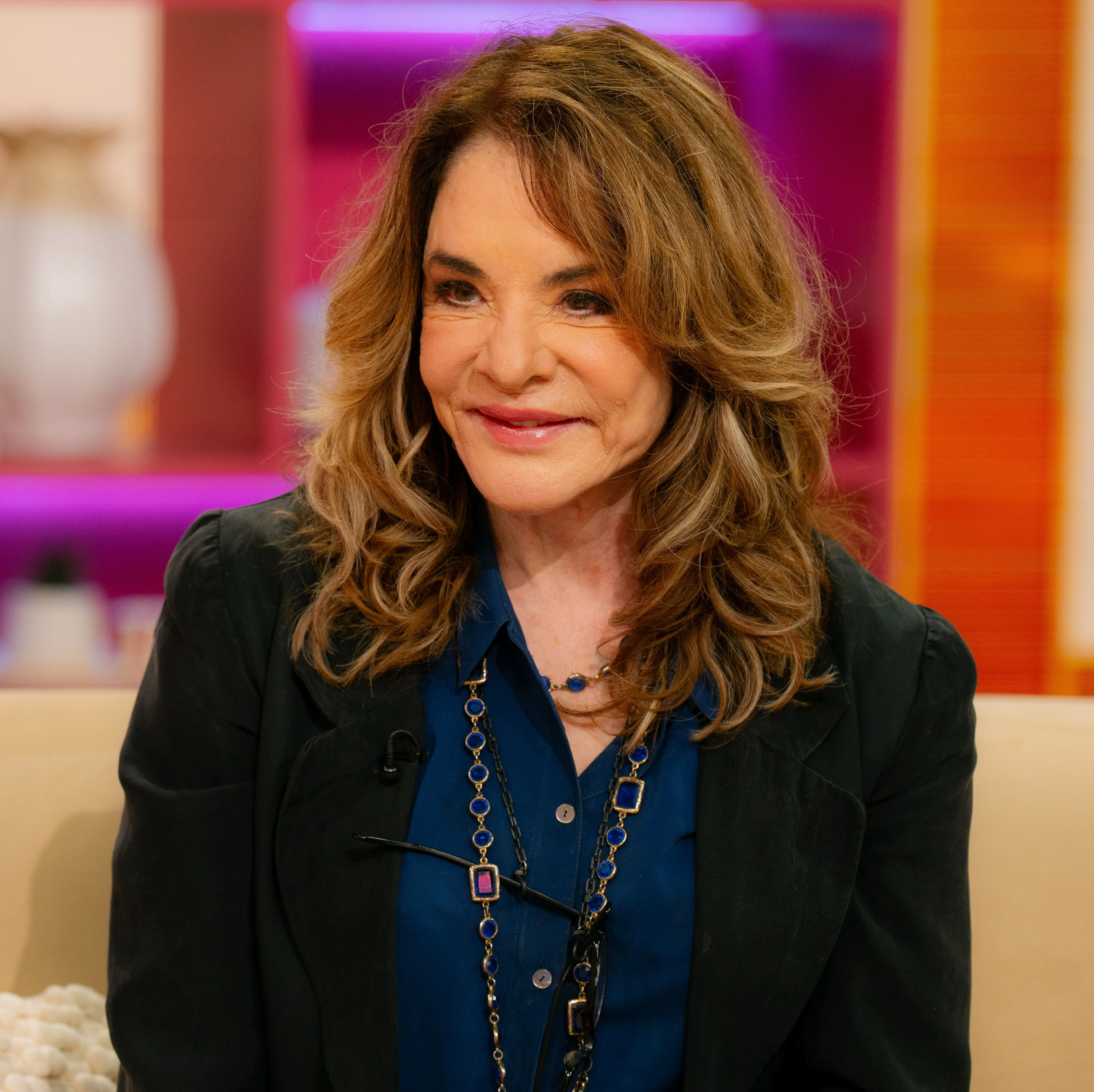 Stockard Channing smiling and looking to her right while wearing a dark blue shirt and a black blazer, with two blue and gold necklaces.