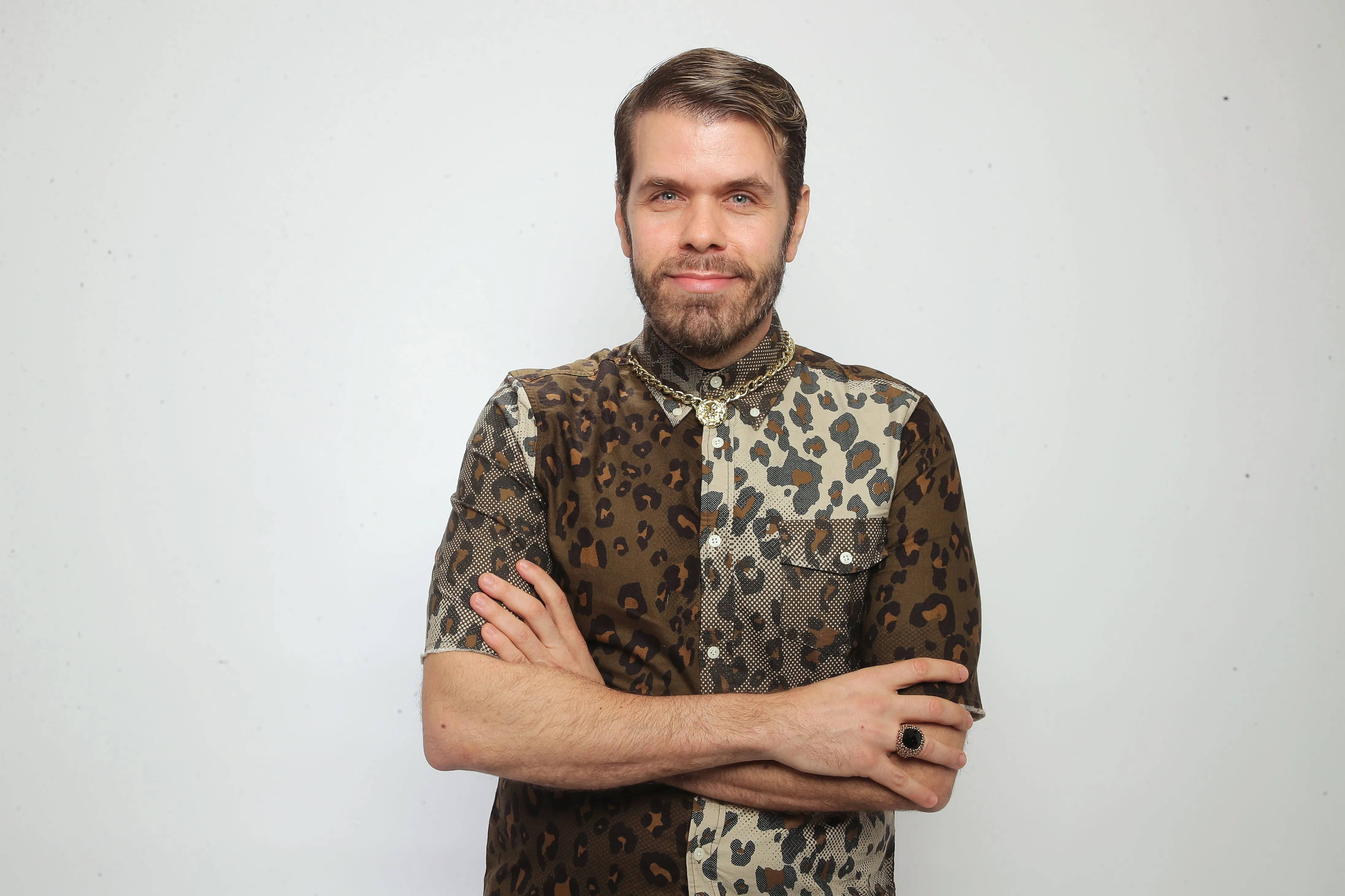 Perez Hilton, a man with a beard and a leopard print shirt, smiles at the camera with his arms crossed.