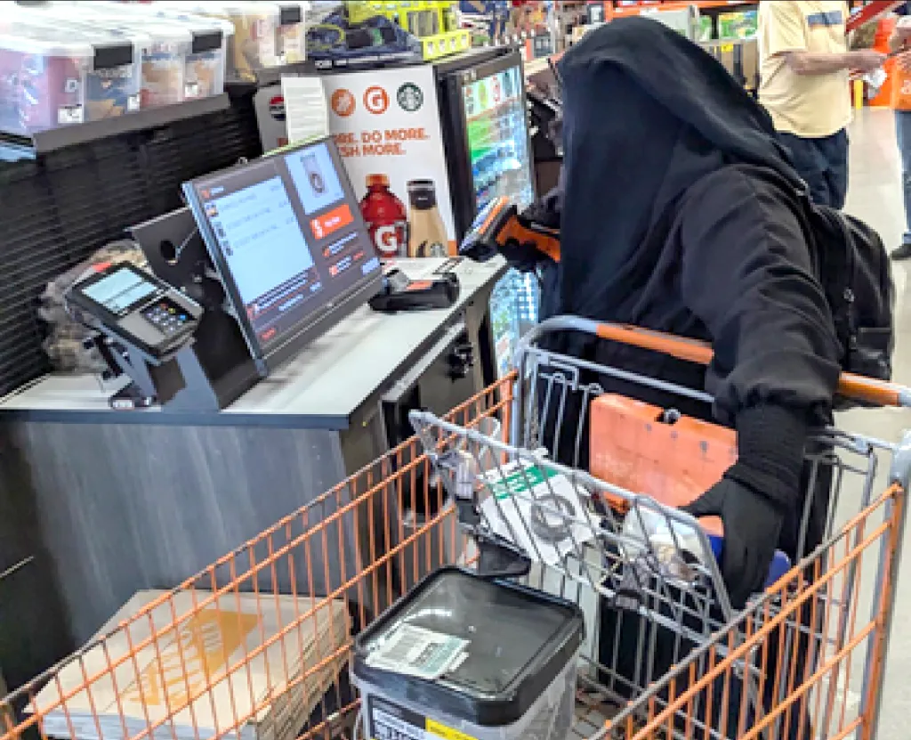 Jessica Bowie in a Home Depot, scanning items at a self-checkout, including a large plastic bin filled with nails in her shopping cart.