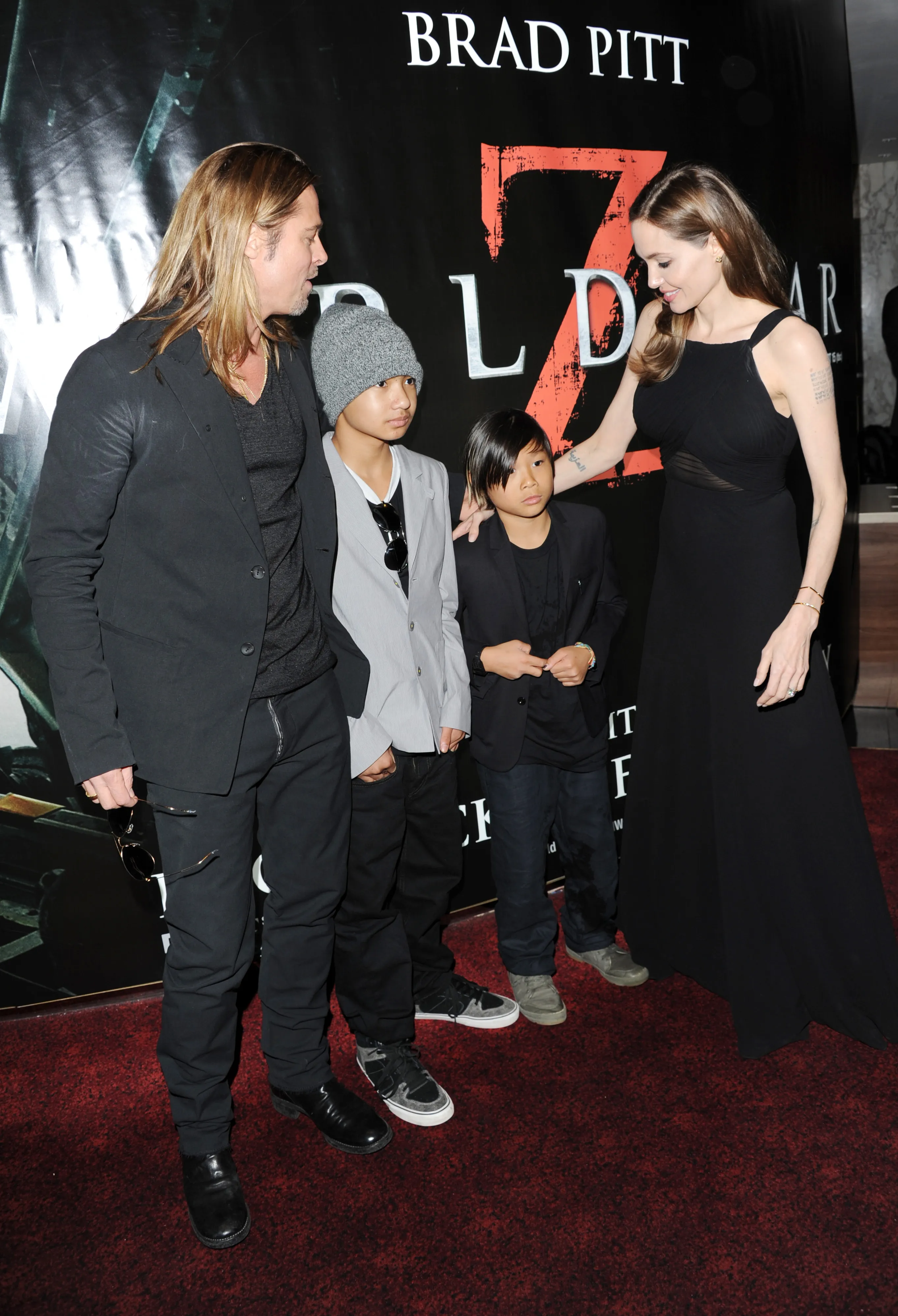 Brad Pitt, Maddox Jolie-Pitt, Pax Jolie-Pitt, and Angelina Jolie standing on a red carpet in front of a movie poster.