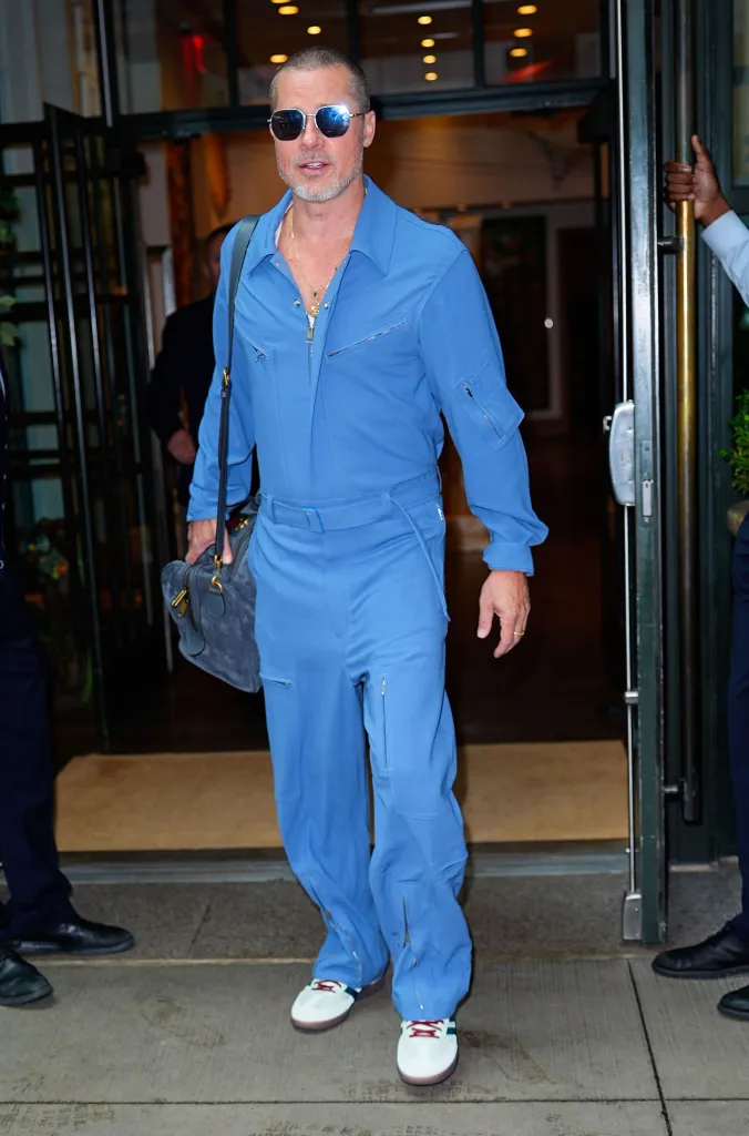 Brad Pitt in a blue jumpsuit, sunglasses, and a gray bag exits a building.