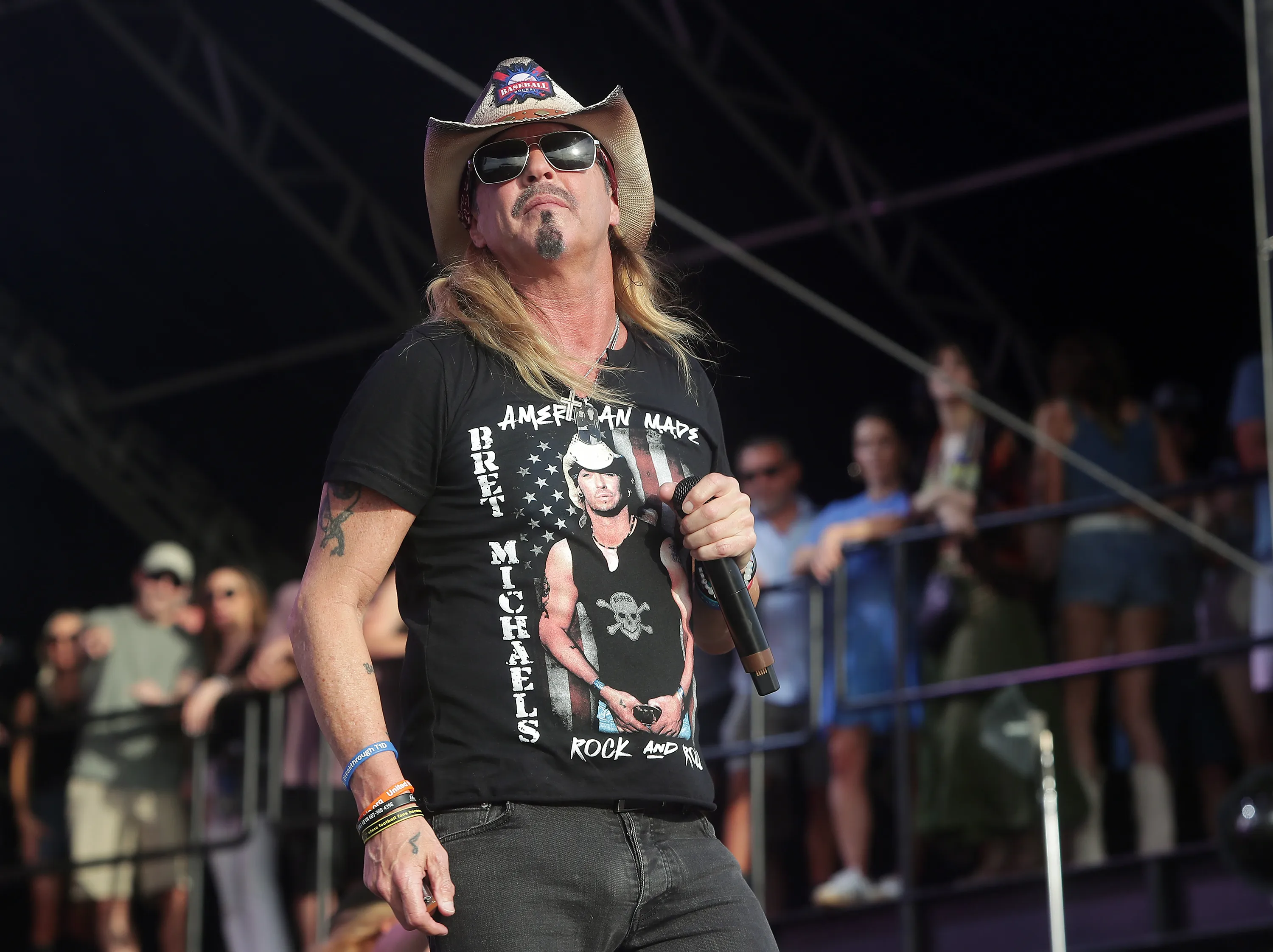 Bret Michaels performing on stage, wearing a cowboy hat, sunglasses, and a black t-shirt featuring his own image.