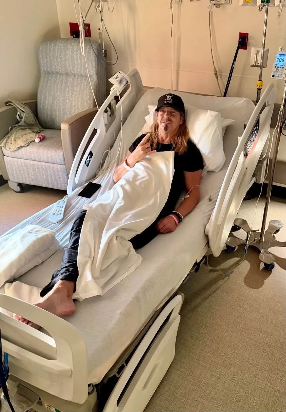 Bret Michaels in a hospital bed making a rock and roll hand gesture.