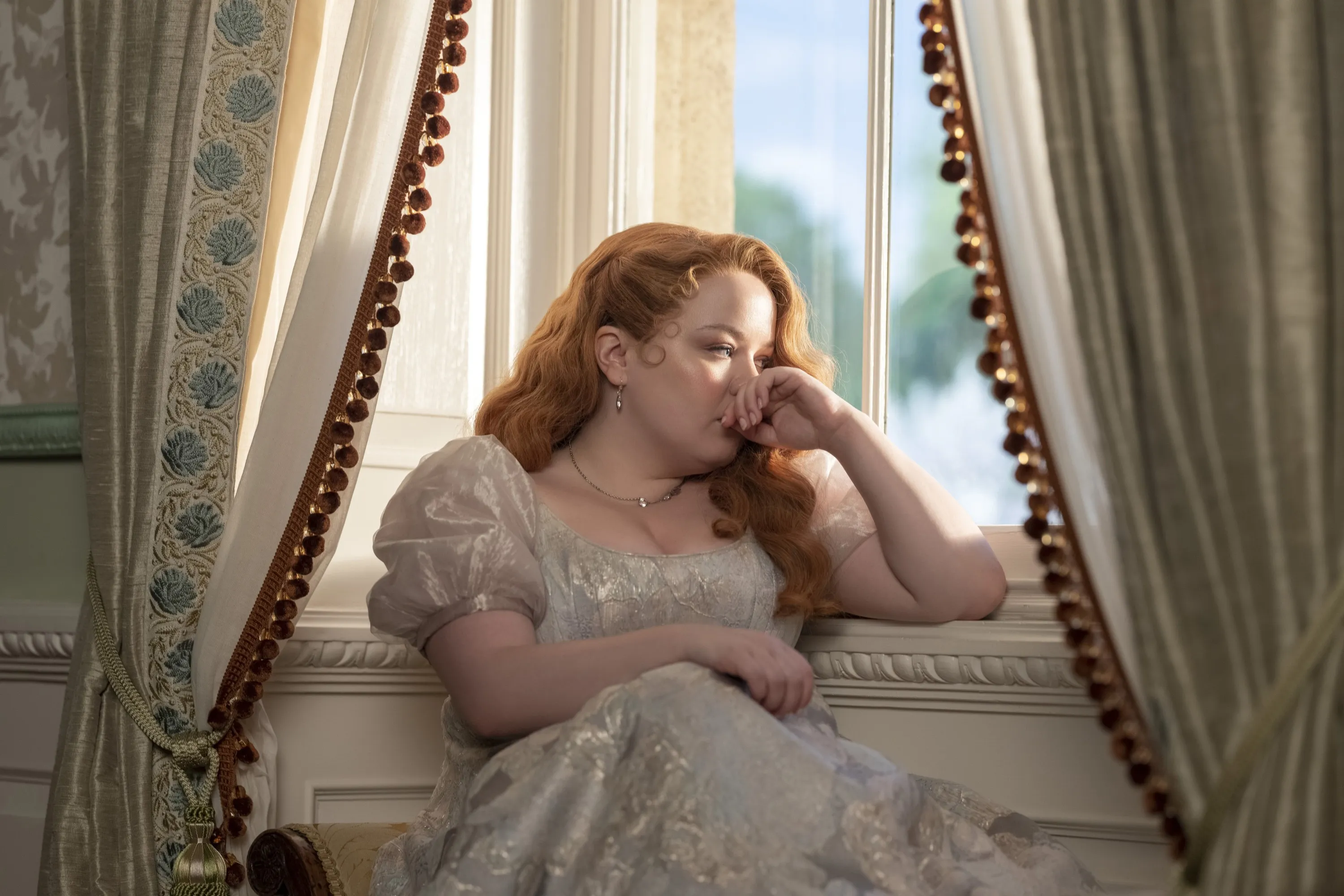 Nicola Coughlan as Penelope Featherington in Season 3 of Bridgerton, sitting by a window, looking sad.