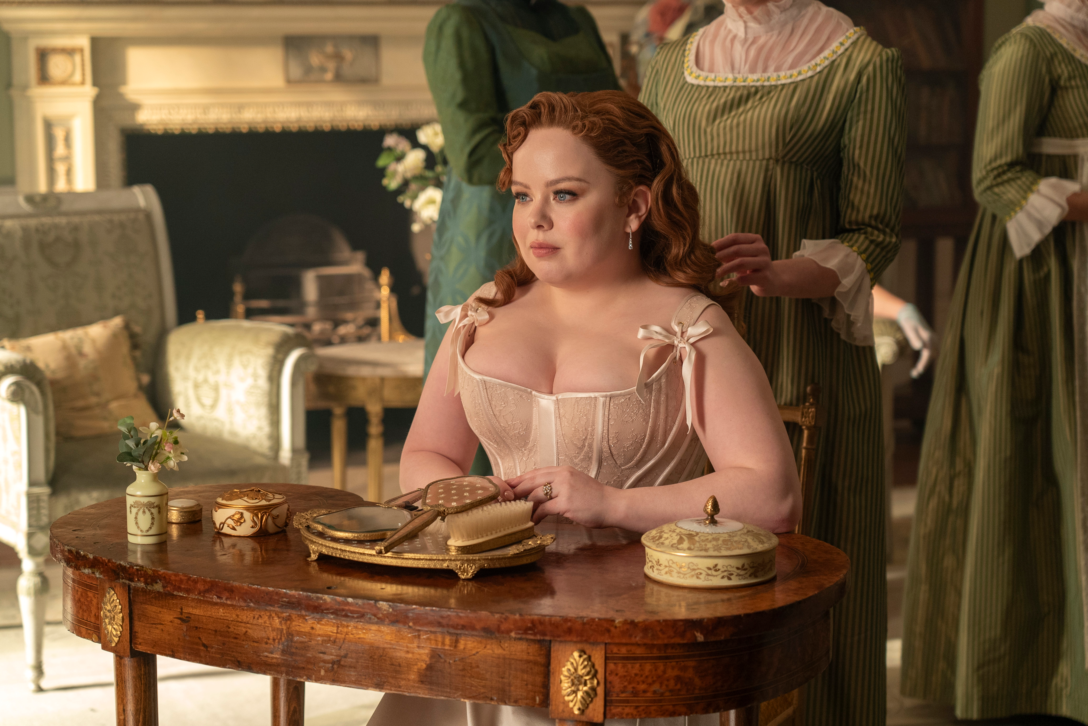 Penelope Featherington in a cream corset and skirt, with a hairbrush and mirror set on the table in front of her.