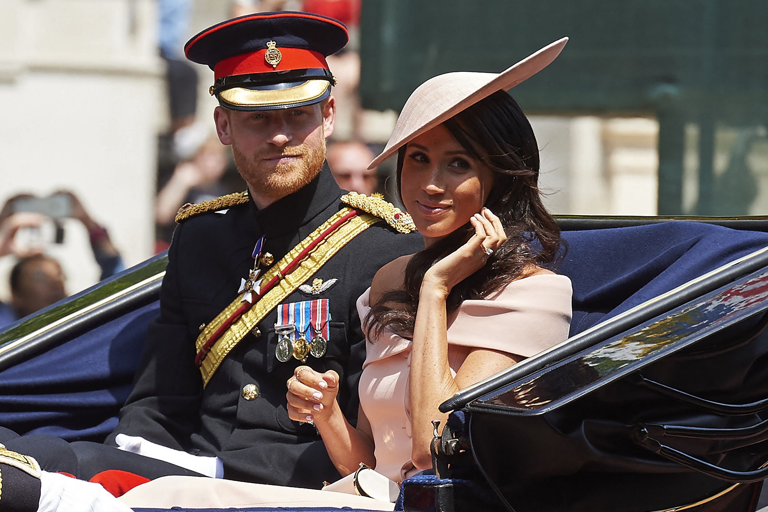 Prince Harry in military uniform and Meghan Markle in a peach dress and hat, riding in a carriage.