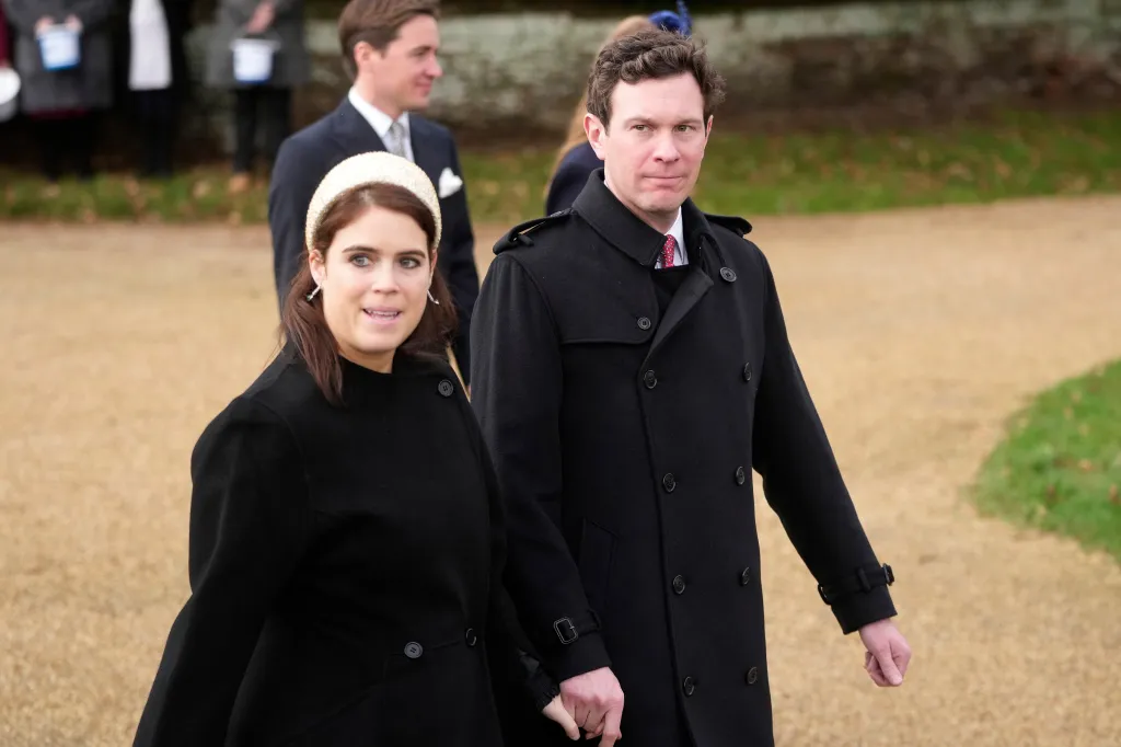 Princess Eugenie, in a black coat and white headband, and Jack Brooksbank, in a black coat, walk hand in hand.