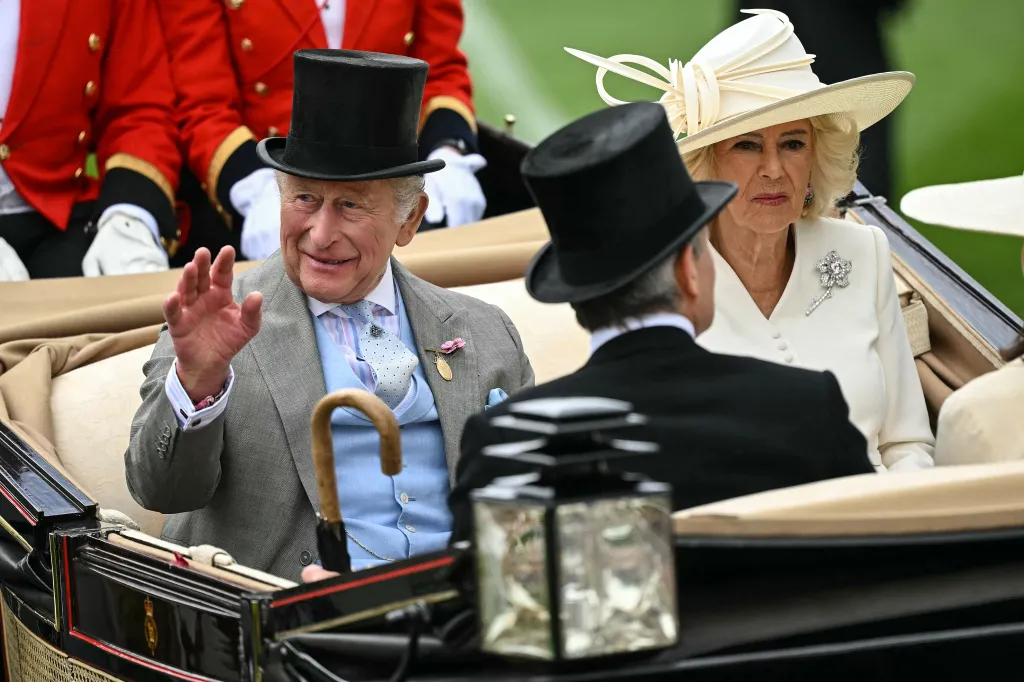 King Charles III, in a grey morning suit and black top hat, waving next to Queen Camilla, in a cream coat and cream hat, at Royal Ascot.