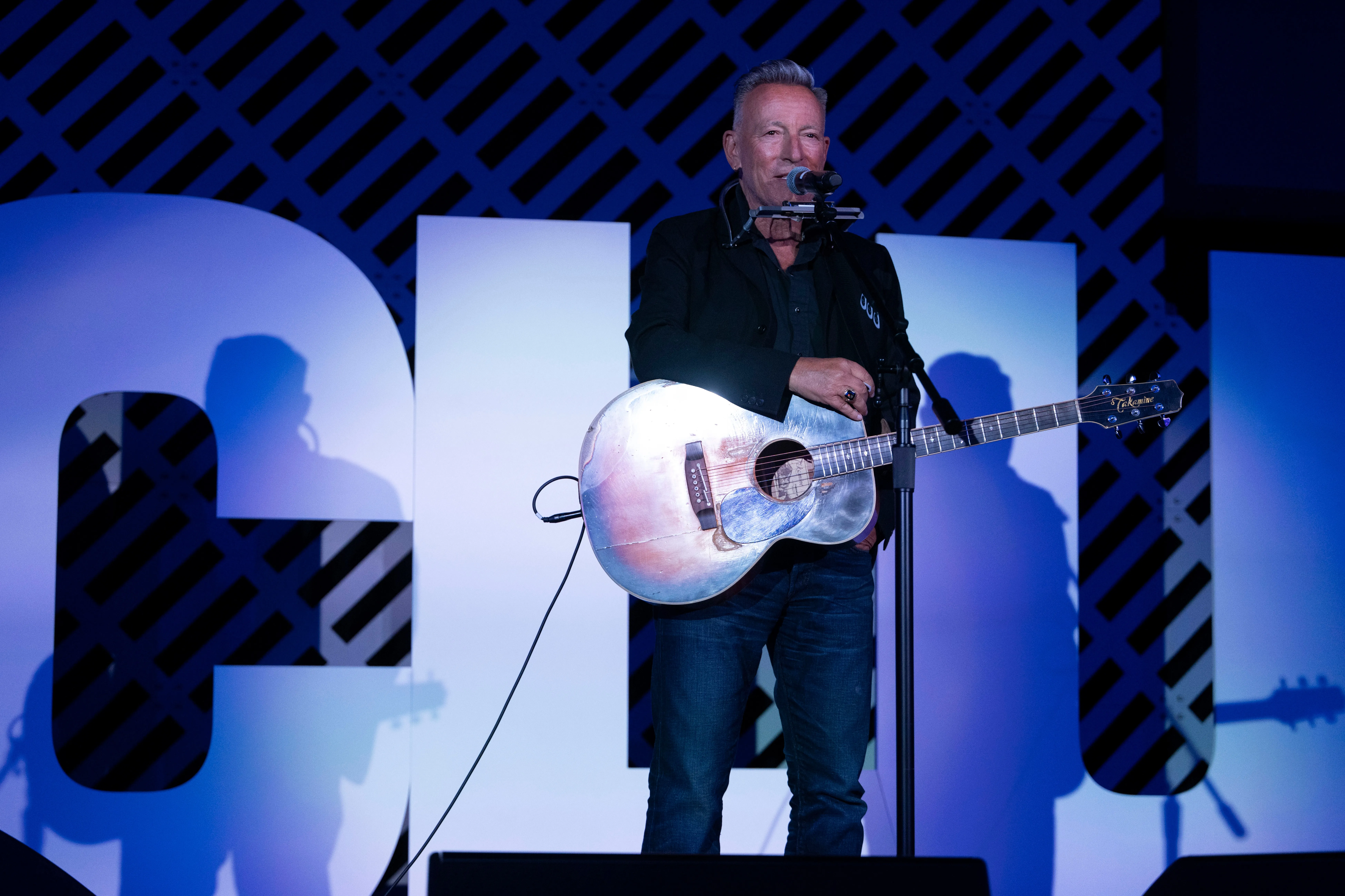 Bruce Springsteen performing on stage with a harmonica around his neck and an acoustic guitar.