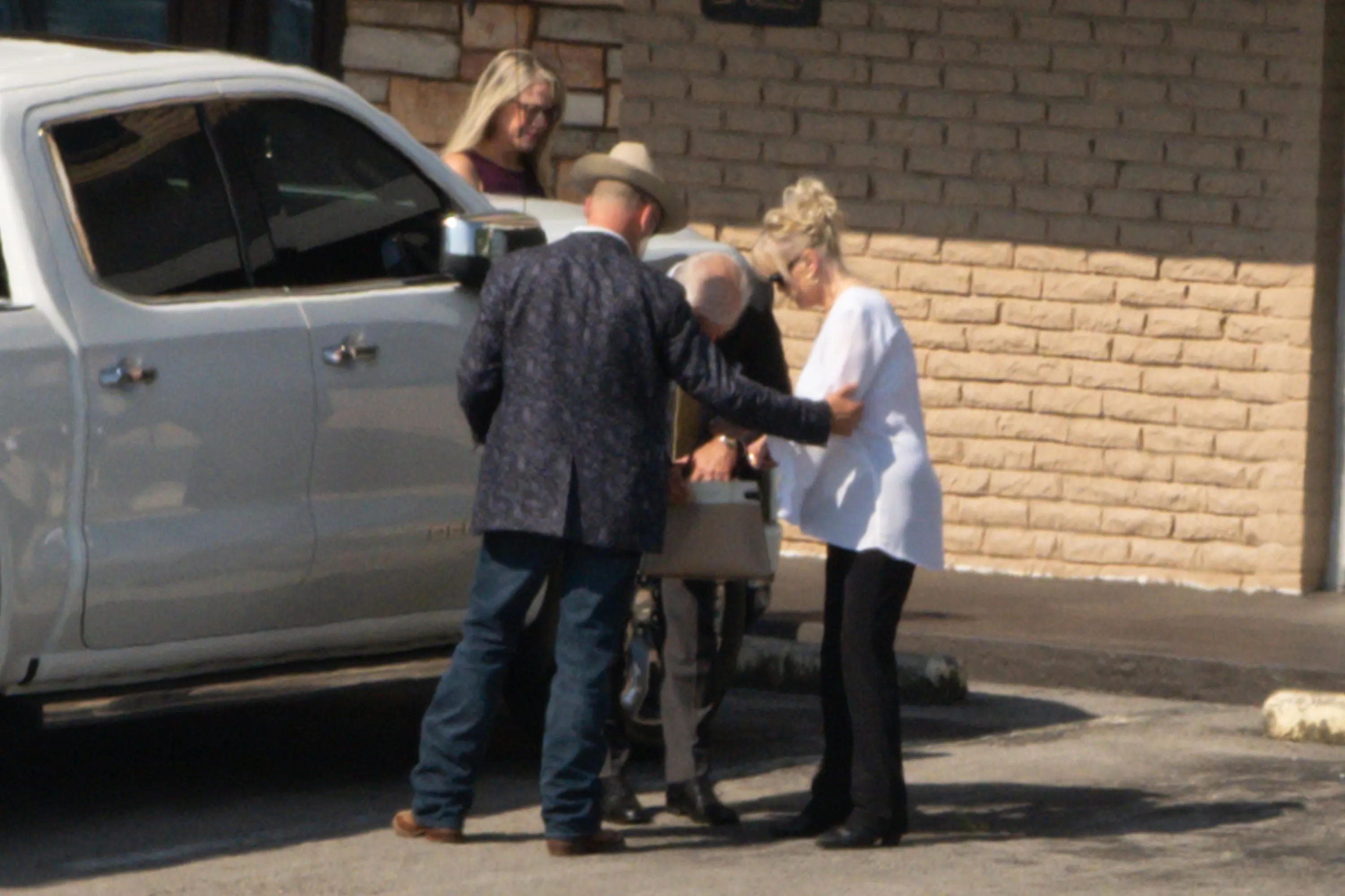 Bryan Seaver, in a patterned jacket and cowboy hat, assists an elderly man and woman next to a white truck with a brick building in the background.