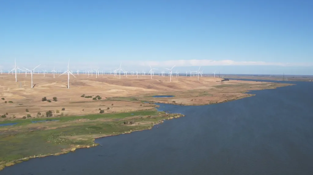 Wind turbines on a tan landscape next to a body of water under a clear blue sky.