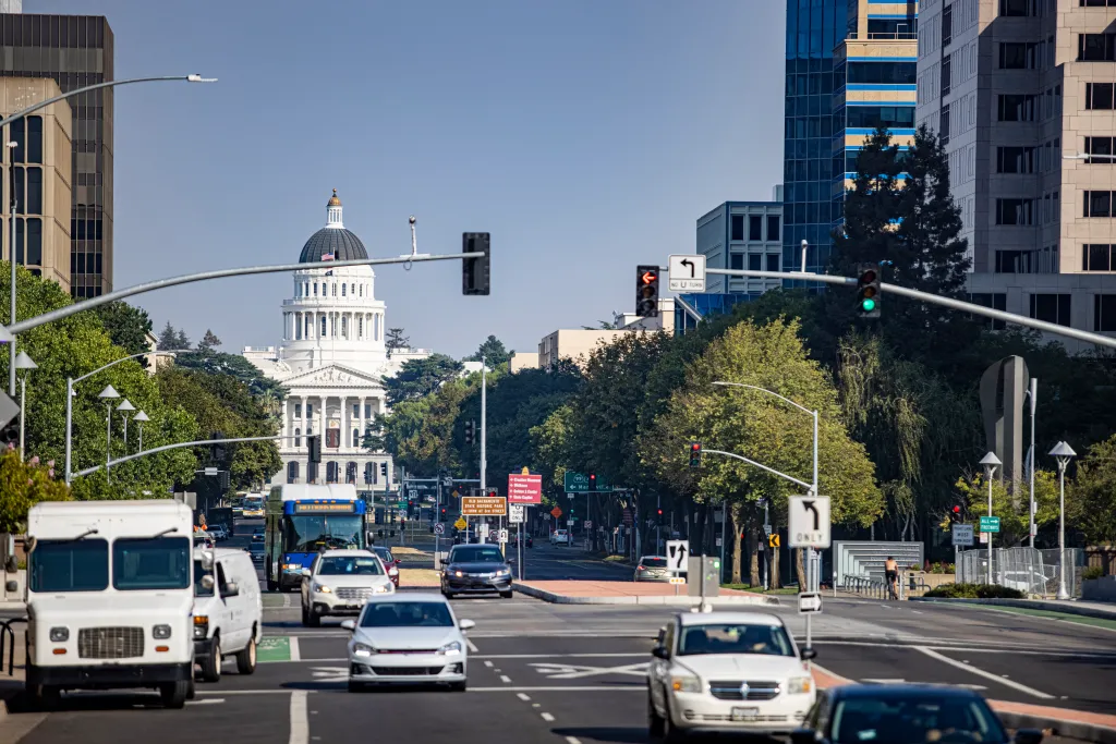 The California State Capitol building is visible in the background, with a busy street full of cars and buses in the foreground.
