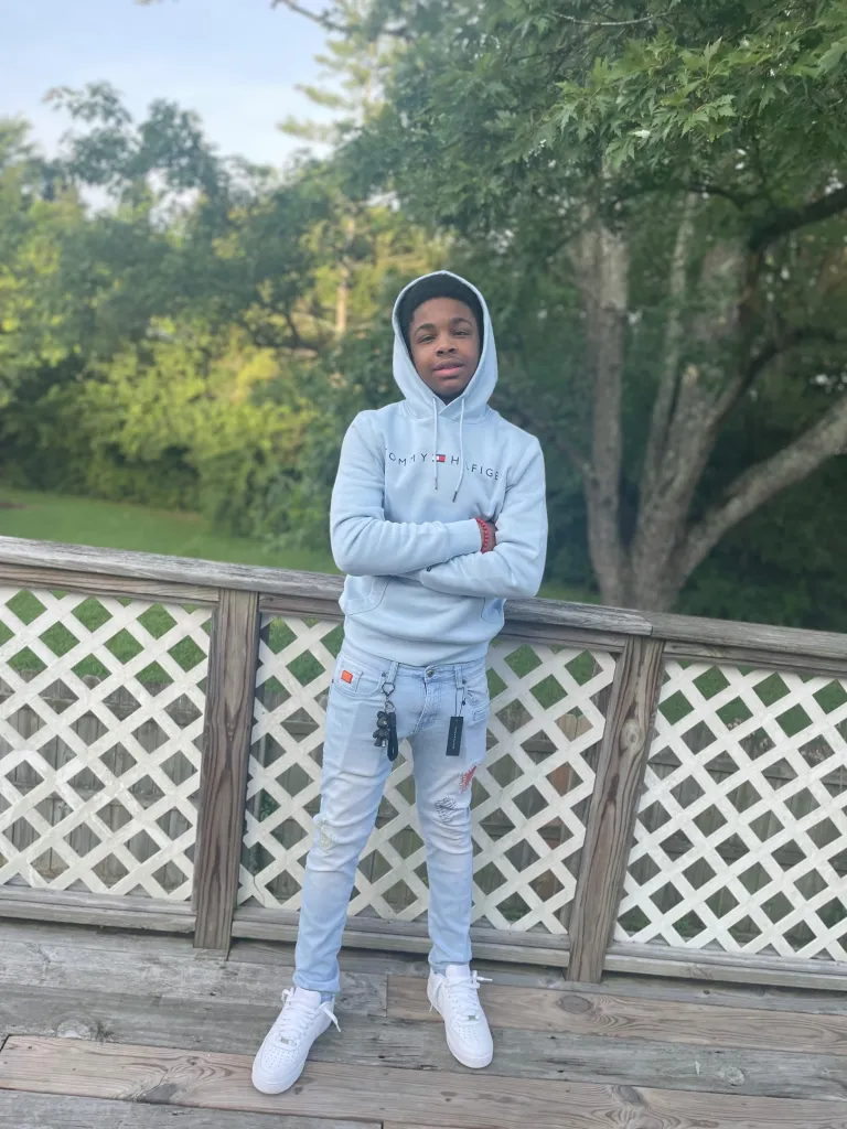 Cameron Frierson, a 16-year-old Black male, stands on a wooden deck with his arms crossed, wearing a light blue Tommy Hilfiger hooded sweatshirt, ripped jeans, and white sneakers.