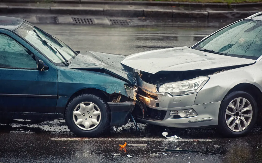Two cars damaged from a head-on collision on a wet road during rain.
