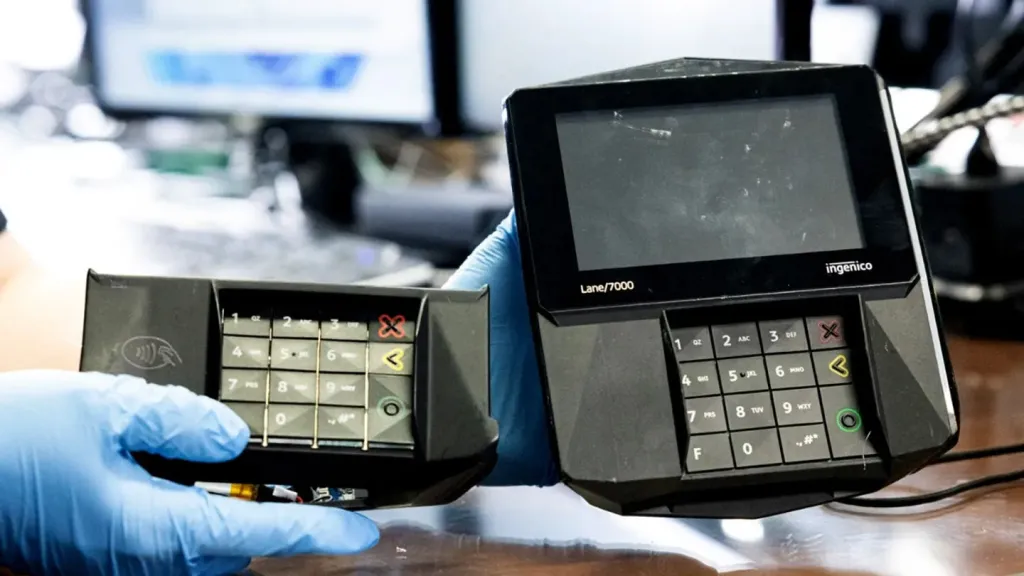 A person in blue gloves holds a card skimmer next to a payment terminal.