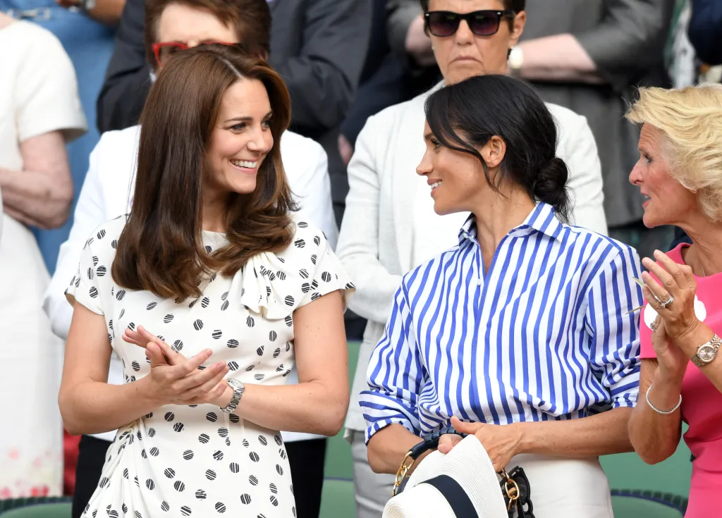 Catherine, Duchess of Cambridge, in a black and white polka dot dress and Meghan, Duchess of Sussex, in a blue and white striped shirt, cream pants and cream hat, attend the Wimbledon Tennis Championships.
