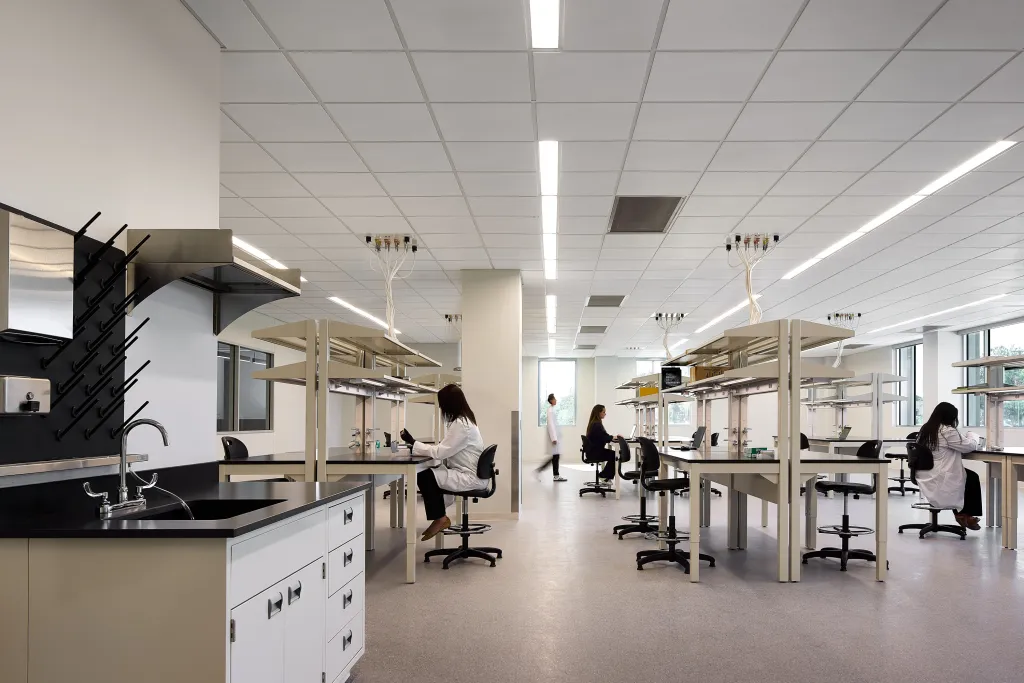 Workers in white lab coats and dark clothes in a large, bright laboratory with several workbenches.