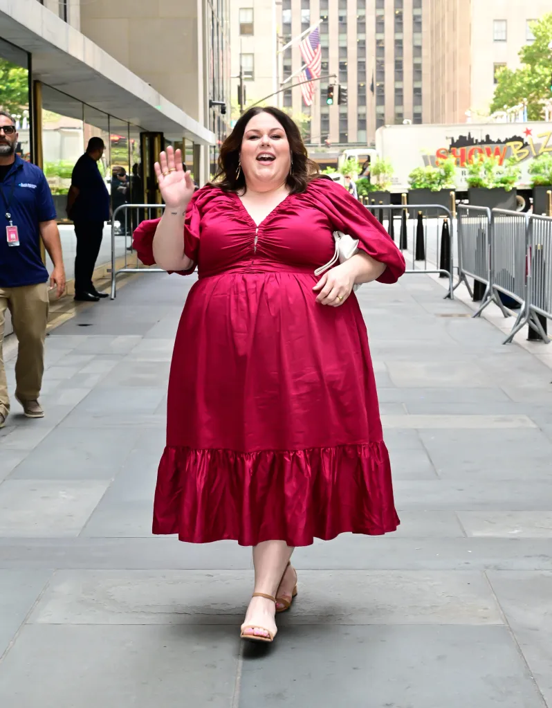 Chrissy Metz smiling and waving while walking outdoors in a magenta dress.