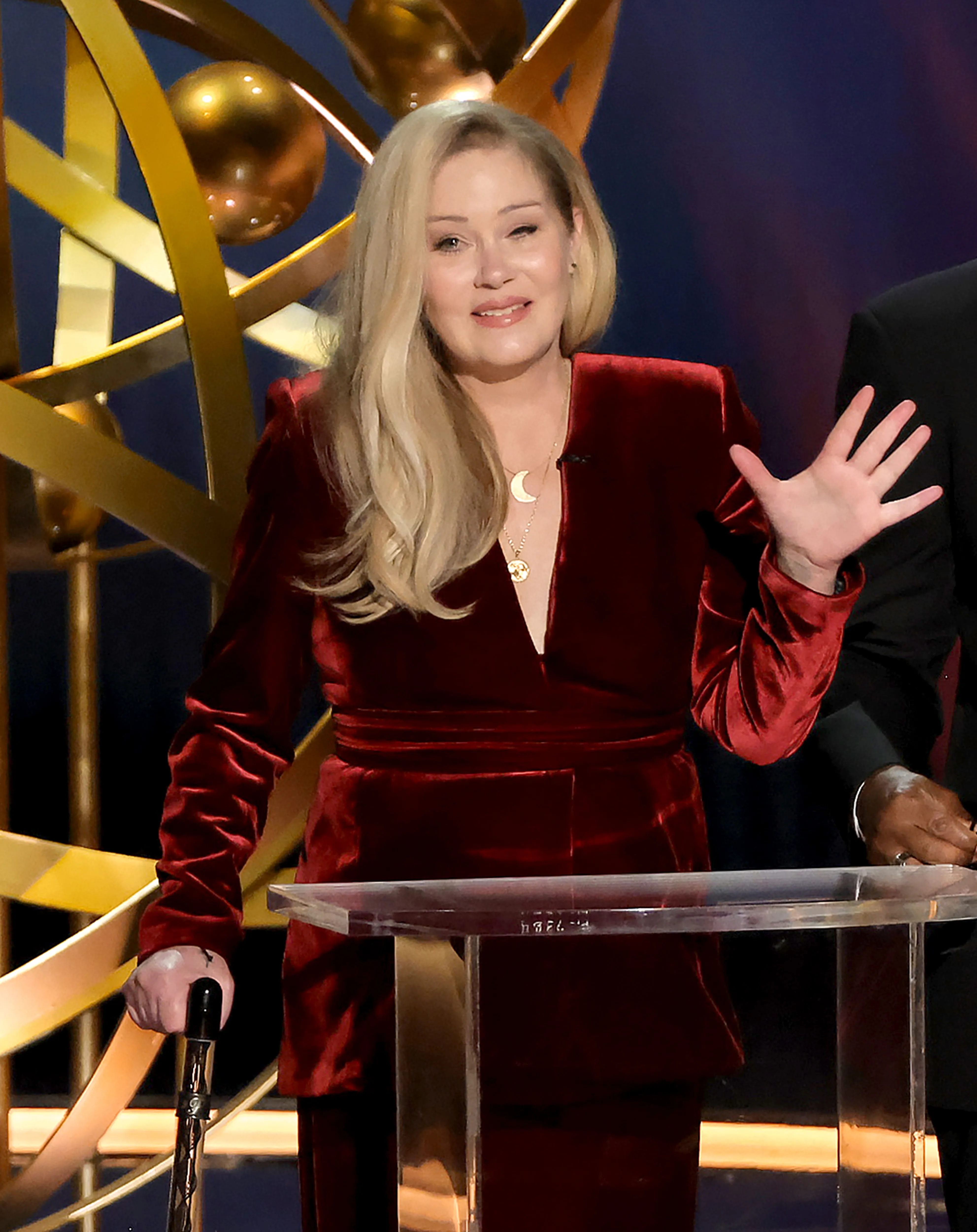 Christina Applegate at a podium, wearing a dark red velvet suit and holding a cane, waving her left hand.
