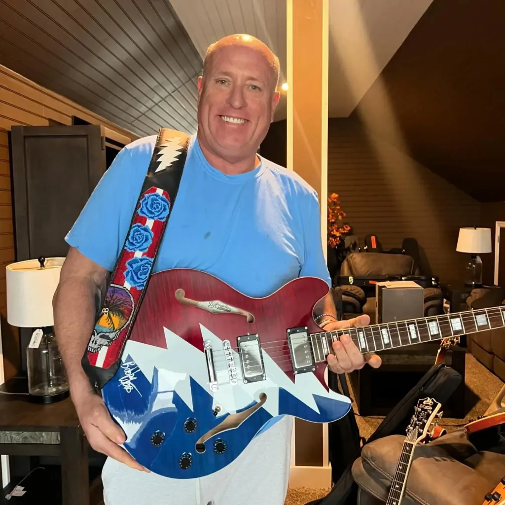 Christopher Durnan smiling while holding a red, white, and blue electric guitar.