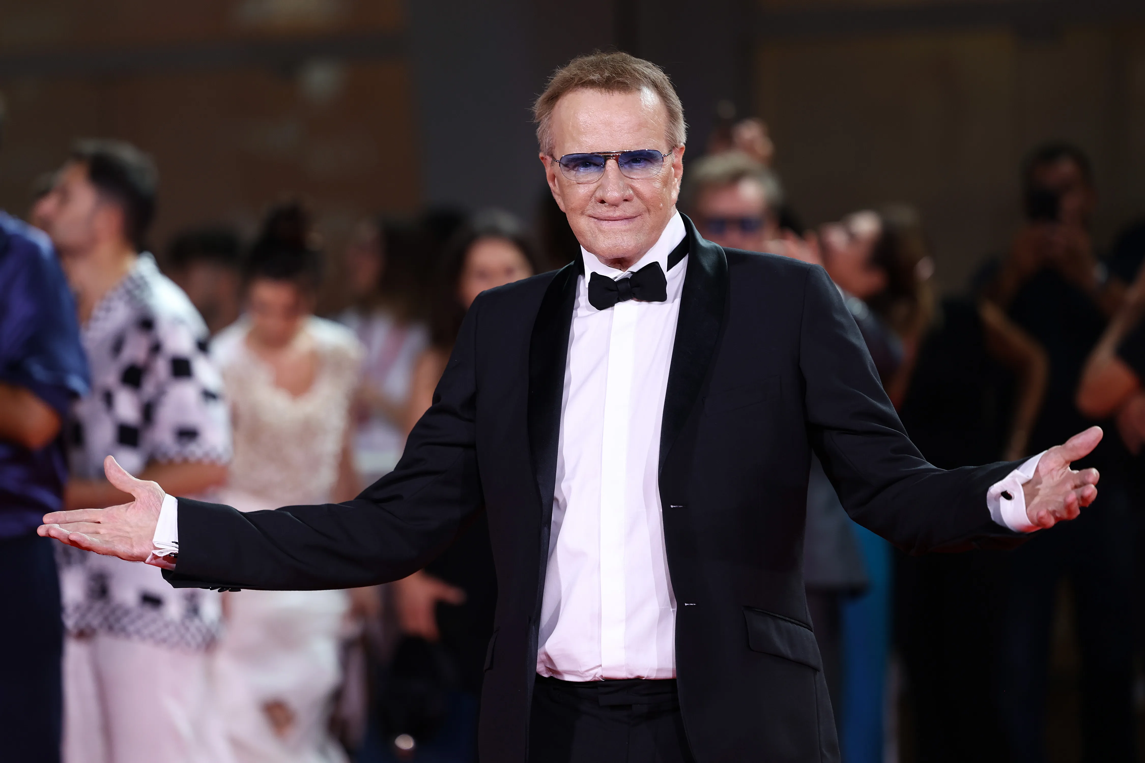 Christopher Lambert smiling on the red carpet, arms outstretched, wearing a tuxedo and blue-tinted glasses.