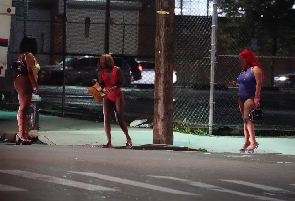 Three women in skimpy clothing and high heels stand on a street at night, with a chain-link fence and parked cars in the background.