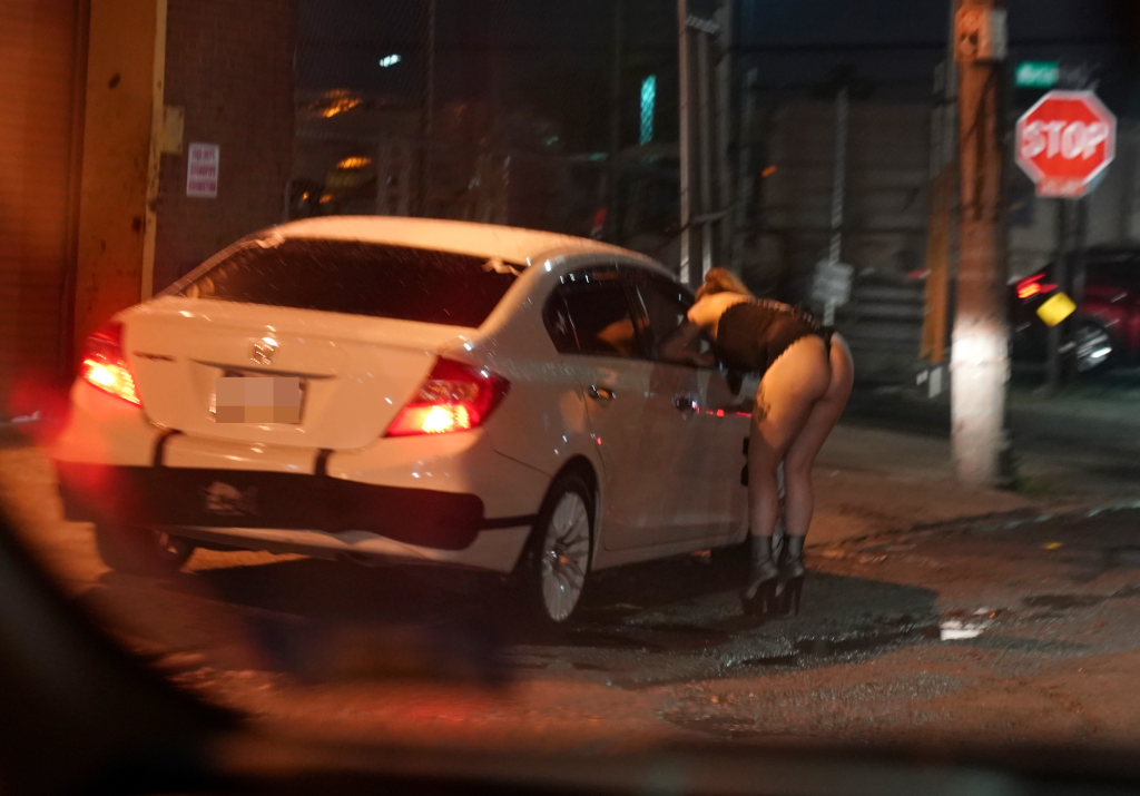 A person wearing high heels and a thong bending into the window of a white car at night.