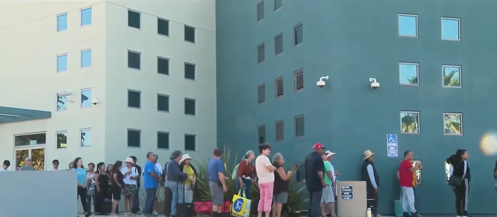 A line of people waiting outside a light-colored building with multiple windows.