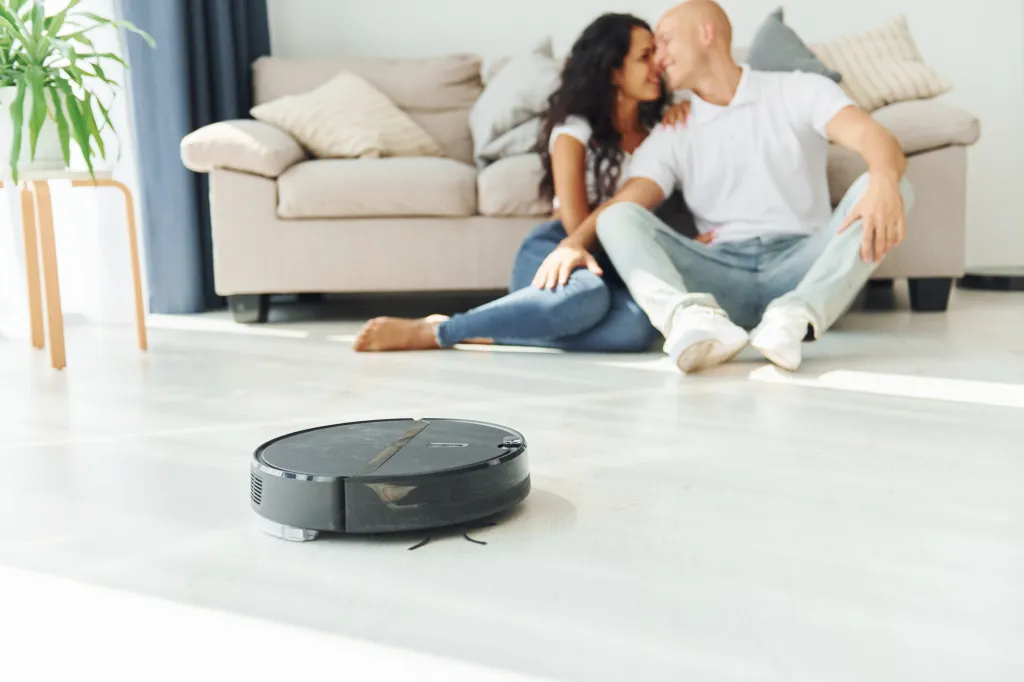A robot vacuum cleaner on a light wooden floor with a couple embracing in the background.