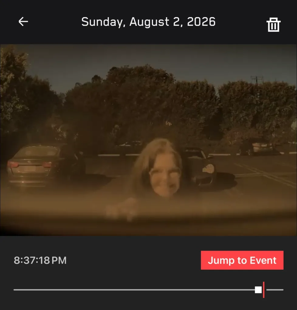 Comedian Laura Kightlinger smiling and writing on a fogged car windshield as captured by a Tesla security camera.
