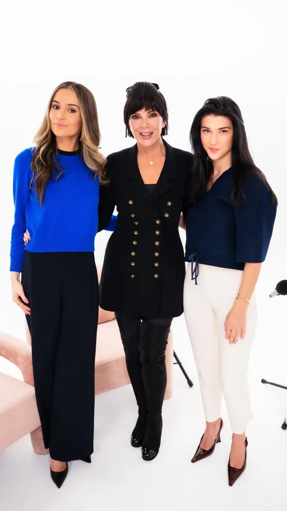 Three women, Kris Jenner in the middle, smiling and standing arm-in-arm.