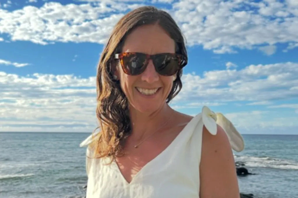A woman with sunglasses smiles in front of the ocean.