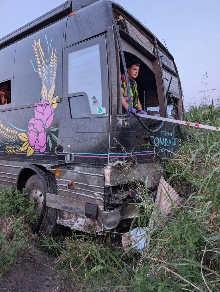 A tour bus, damaged from a crash, with a man looking out from inside.