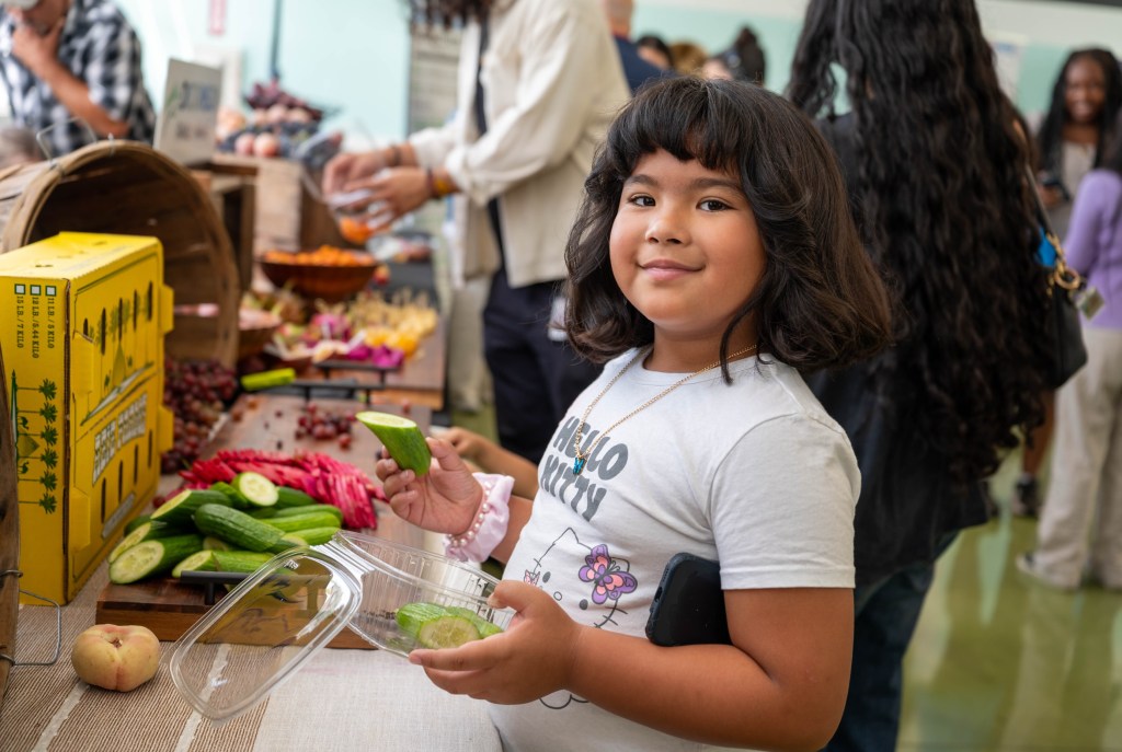 A young girl holding a cucumber slice and a plastic container, smiling at an event featuring student-created menus.