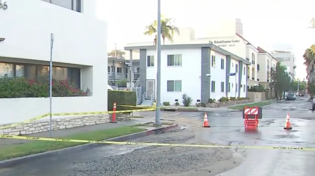 Yellow caution tape blocks a street where water from a broken pipe is flowing across the road.