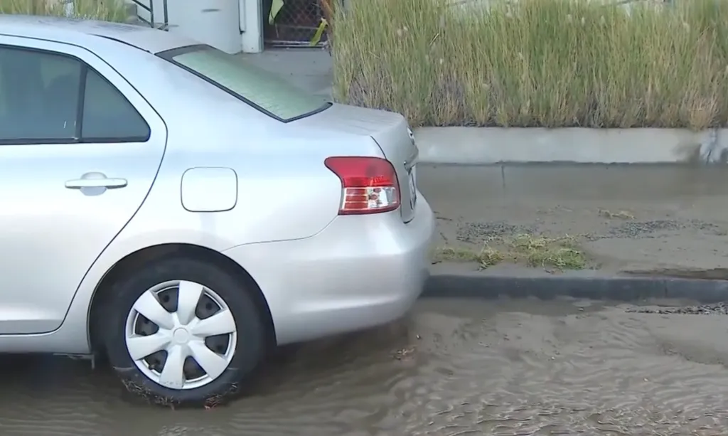 A silver sedan parked on a flooded street, with water partially covering its tires.