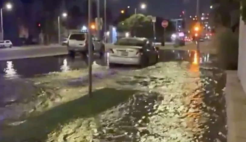 Nighttime street flooding in a residential area of Beverly Grove, with cars partially submerged and blue lights reflecting on the water.